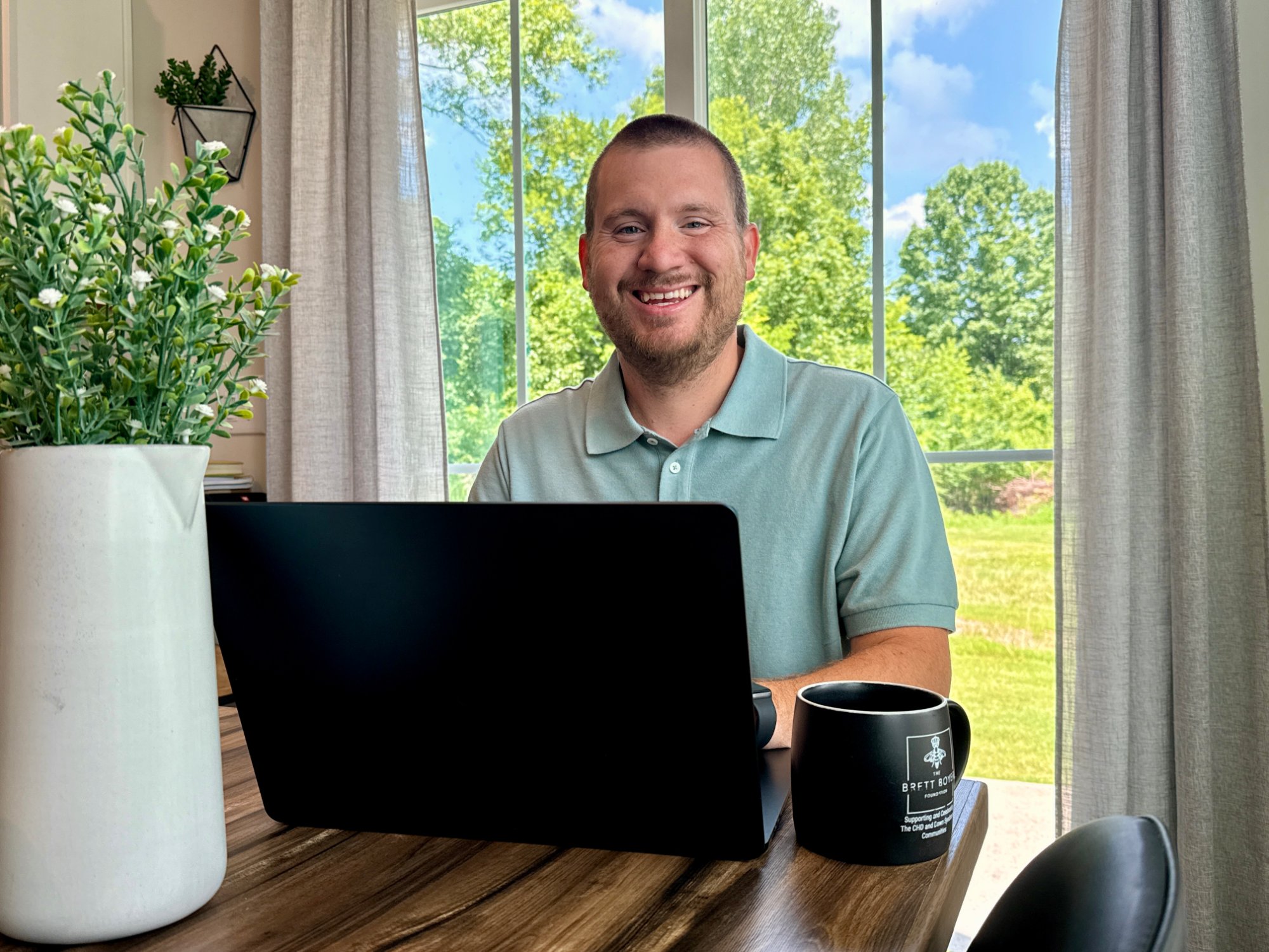 Sean smiling behind his laptop at the dining table with sunlit green trees through the windows behind him